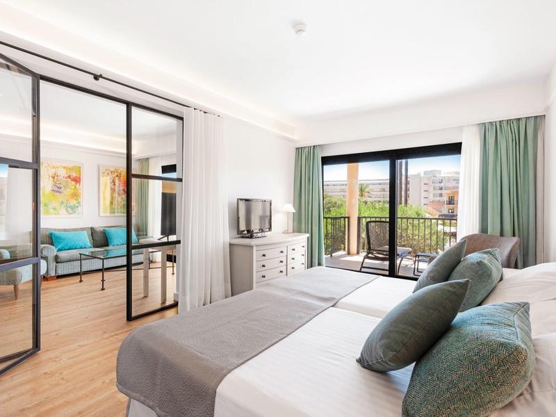 Lichte hotelkamer met tweepersoonsbed, groene kussens, schuifdeuren en balkonzicht.