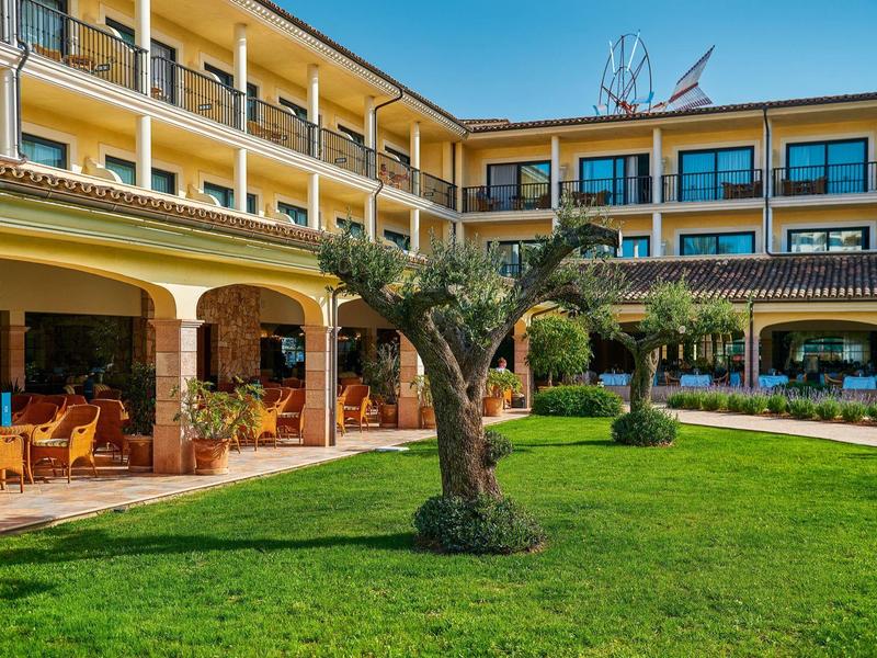Hotelbinnenplaats met groen grasveld, olijboom en balkons aan een geel gebouw.