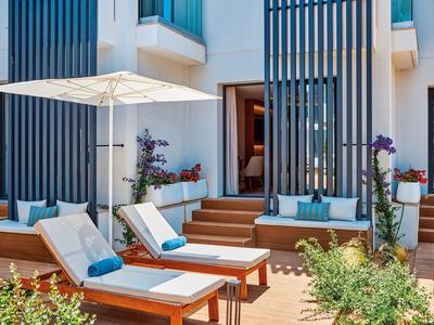 Outdoor lounge area with cushioned wooden sunbeds, umbrella, and plants in front of modern building.