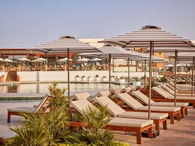 Row of sunbeds with striped umbrellas by a pool at a resort under clear sky.