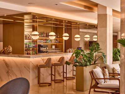 Elegant hotel bar area with marble counter, wooden stools, modern lighting, and cozy seating with plants.