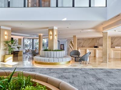 Bright hotel lobby with modern seating, marble floor, plants, and reception area.