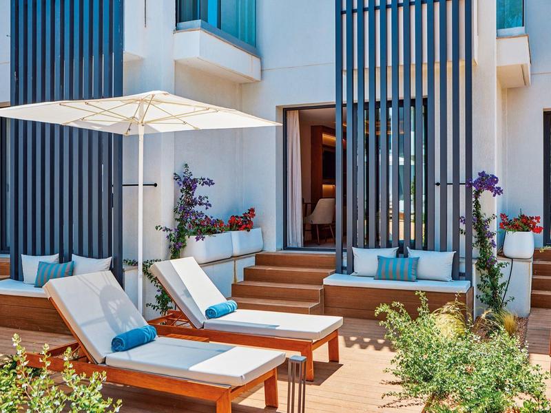 Outdoor lounge area with cushioned wooden sunbeds, umbrella, and plants in front of modern building.