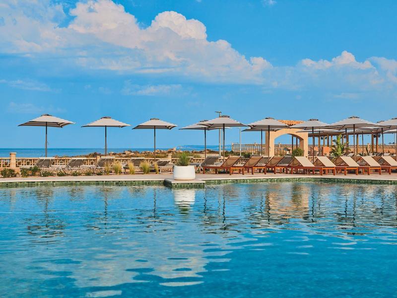 Swimming pool by the sea with sun loungers and umbrellas under a blue sky.