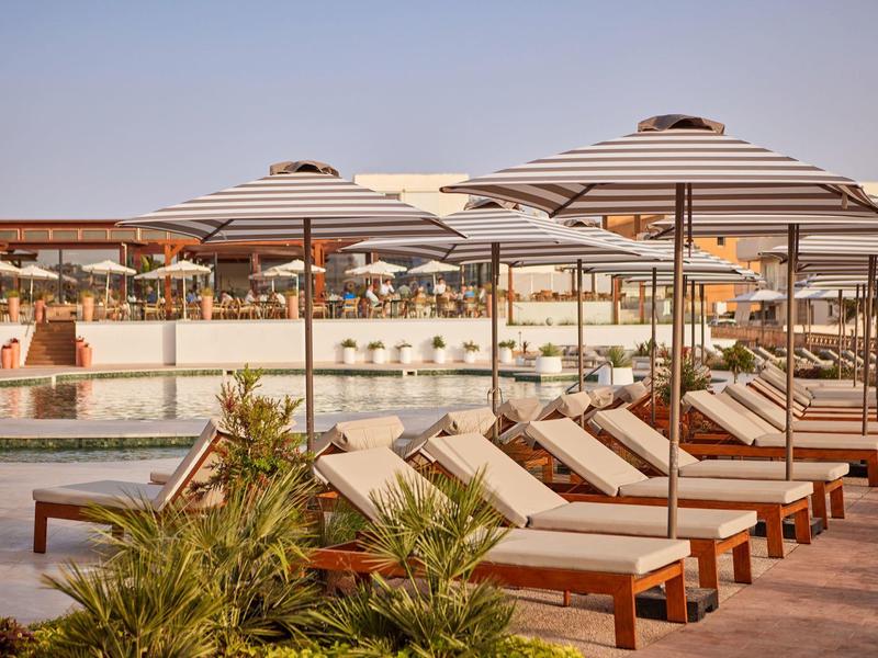 Row of sunbeds with striped umbrellas by a pool at a resort under clear sky.