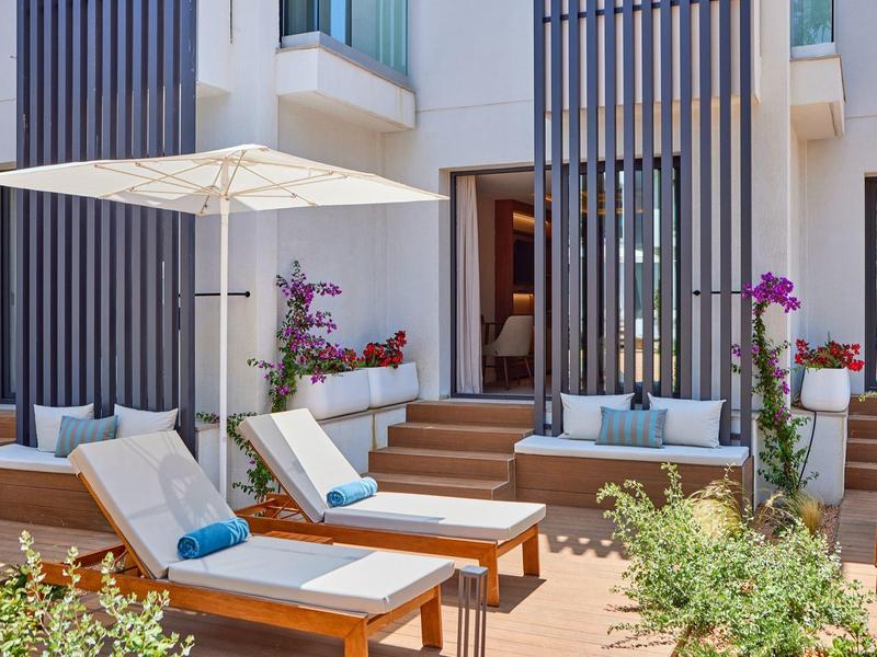 Outdoor lounge area with wooden sunbeds, cushions, umbrella, and open entrance to a room.