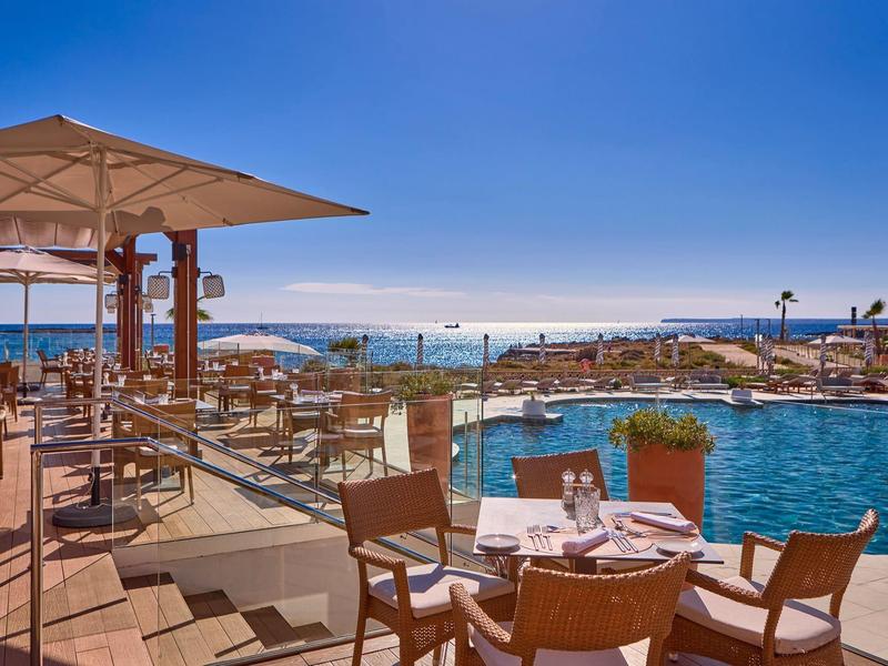 Outdoor hotel dining area beside pool with ocean views under clear blue sky.