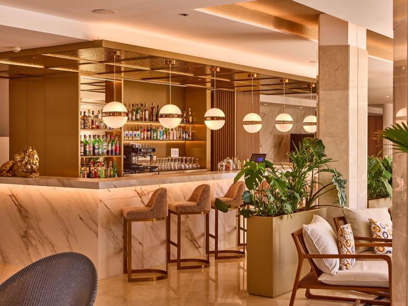 Elegant hotel bar area with marble counter, wooden stools, modern lighting, and cozy seating with plants.
