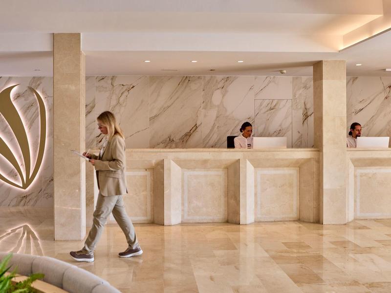 Modern hotel lobby with marble reception desk, three staff, and a walking guest.