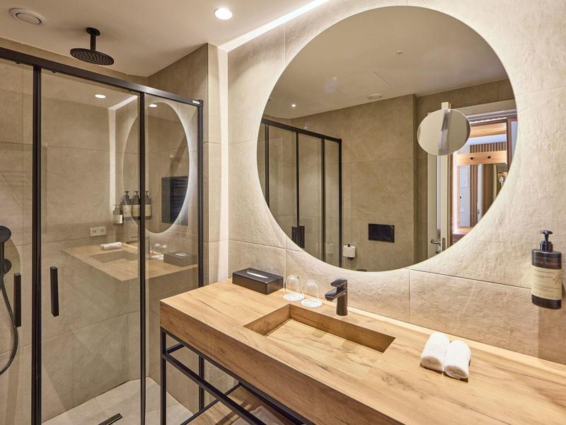 Modern hotel bathroom with wooden vanity, round mirror, and glass shower enclosure.