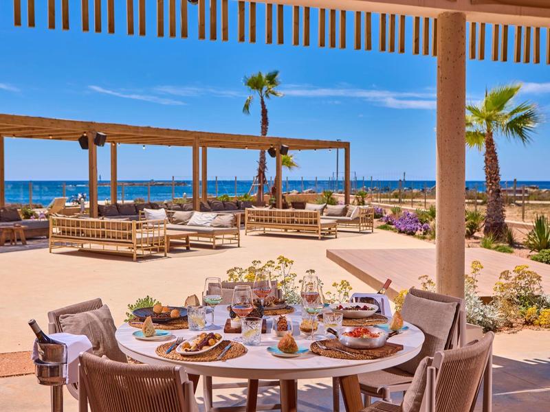 Outdoor dining area with sea view, tables set for meals, sunny skies, and palm trees.