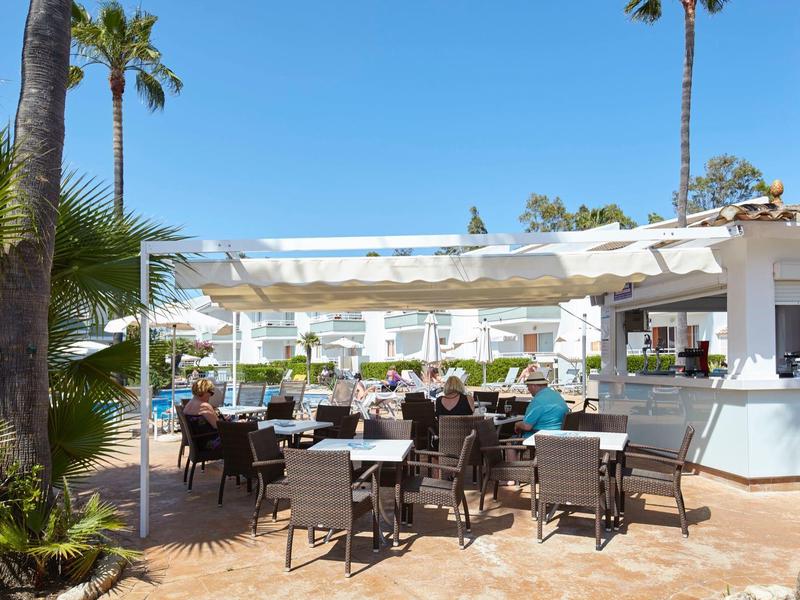 Area esterna di un ristorante con posti a sedere sotto una copertura al sole con cielo blu.