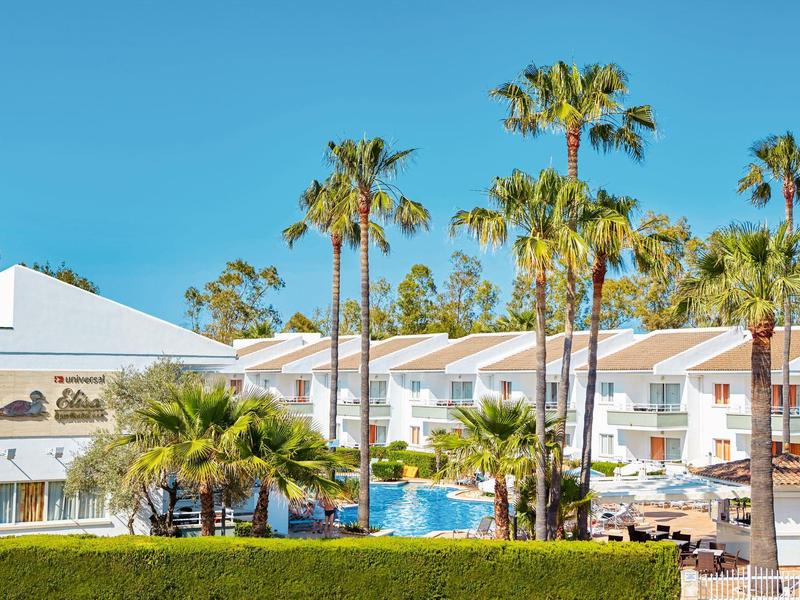 Hotel con piscina, palme e piante fiorite sotto un cielo azzurro limpido.