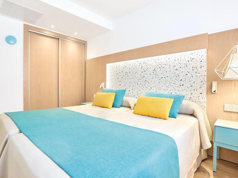 Camera d'hotel moderna con copriletto blu, cuscini gialli e armadio in legno.