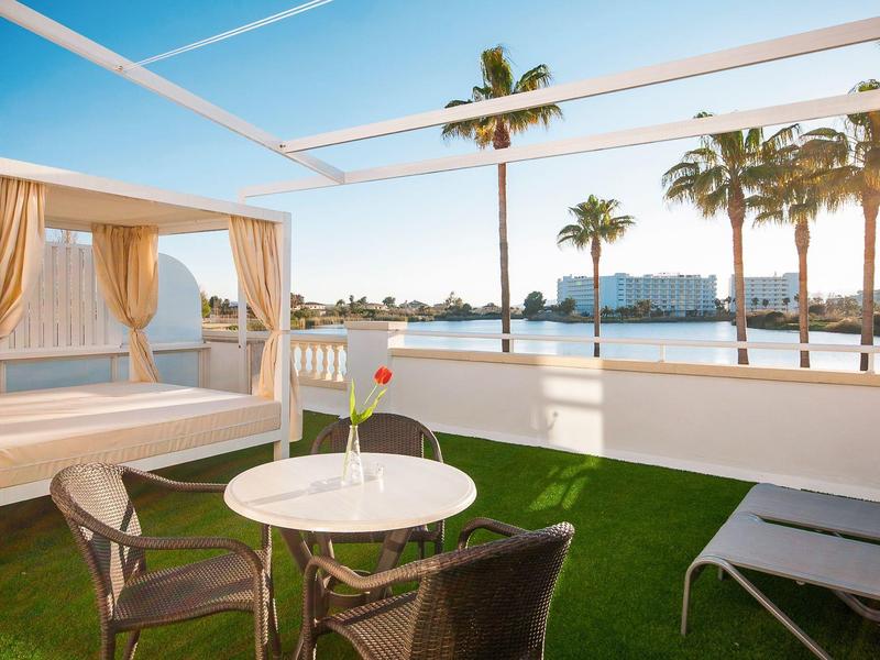 Terrazza con tavolo, sedie e letto da giorno con vista su palme e acqua sotto un cielo limpido.