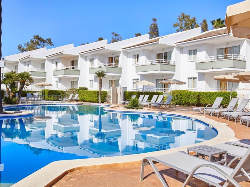 Complesso alberghiero moderno con piscina, edifici bianchi e sedie a sdraio sotto un cielo sereno.