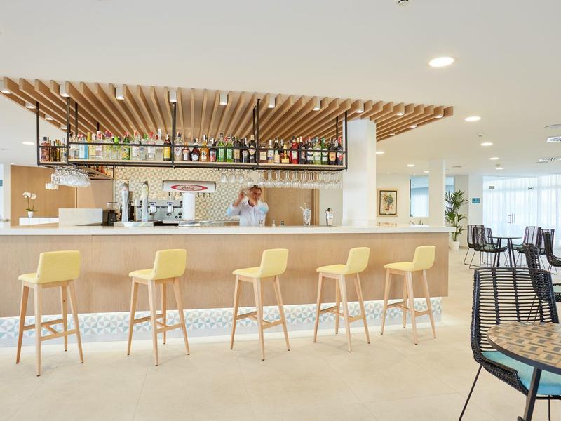 Bar moderno con sgabelli gialli e scaffali di bottiglie in un ambiente luminoso.