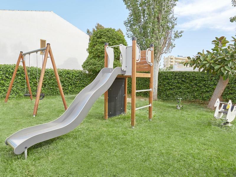 Area gioco per bambini con scivolo, altalena e dondolo su prato verde vicino a una siepe.