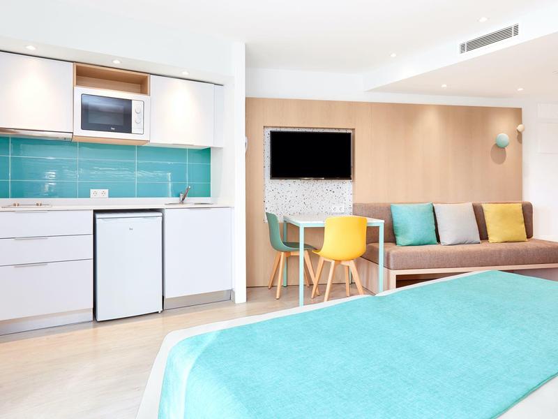 Camera d'hotel moderna con angolo cottura, tavolo da pranzo, divano e letto con coperta turchese.