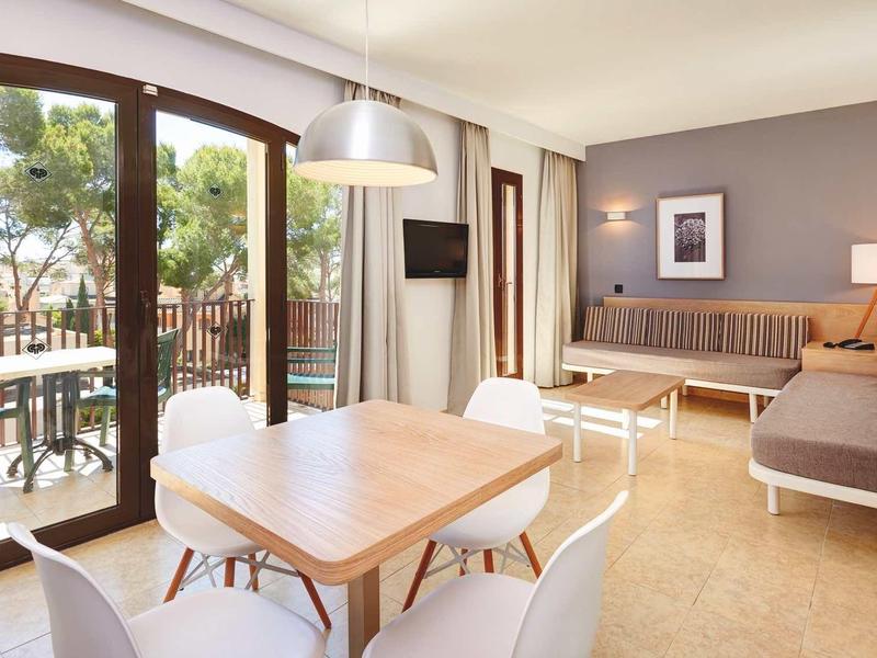 Jasna sala z drewnianym stołem, białymi krzesłami i dużym oknem balkonowym.