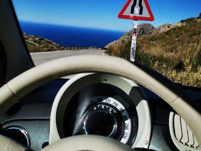 Blick aus einem Auto auf eine kurvige Straße mit einem Verkehrszeichen vor blauem Himmel und Meer.