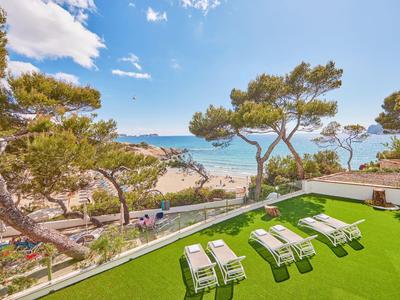 Blick auf Strand mit blauem Himmel, Pinien, Rasenfläche und weißen Liegestühlen im Vordergrund.