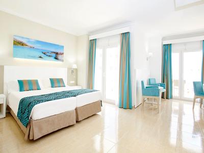 Jasny pokój hotelowy z łóżkiem podwójnym, turkusowymi akcentami i dużymi oknami.