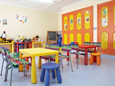 Pokój zabaw dla dzieci z kolorowymi stołami, krzesłami i żółto-pomarańczowymi szafkami.