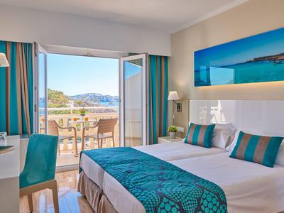 Jasny pokój hotelowy z podwójnym łóżkiem, niebiesko-zielonym wystrojem i widokiem na taras i morze.