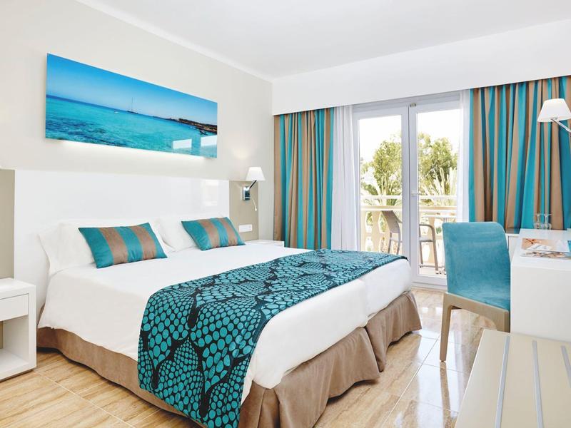 Jasny pokój hotelowy z podwójnym łóżkiem, turkusowymi akcentami i widokiem na balkon.