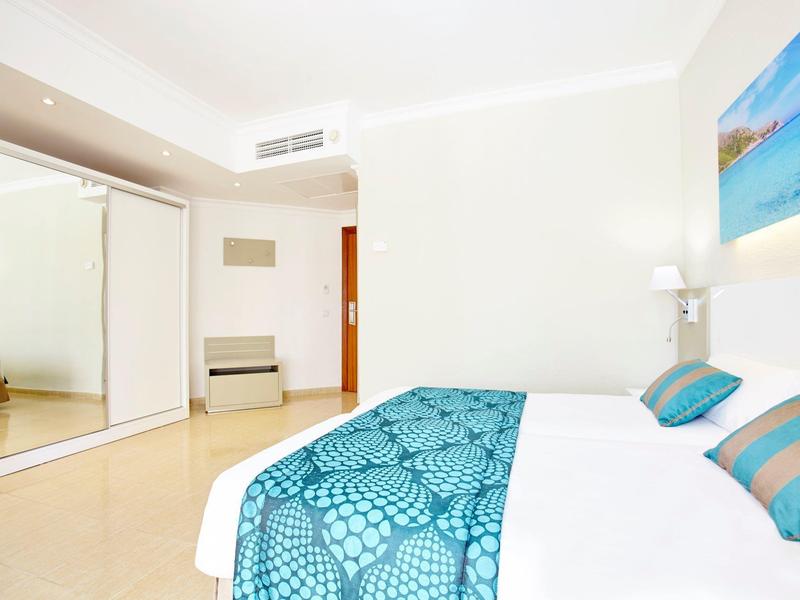 Jasny pokój hotelowy z dużym łóżkiem, turkusowymi poduszkami i szafą z lustrem.