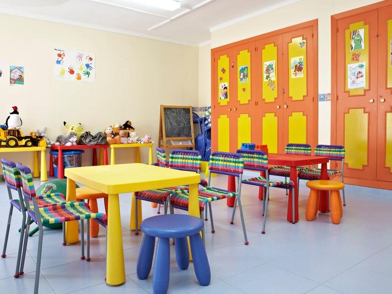 Pokój zabaw dla dzieci z kolorowymi stołami, krzesłami i żółto-pomarańczowymi szafkami.