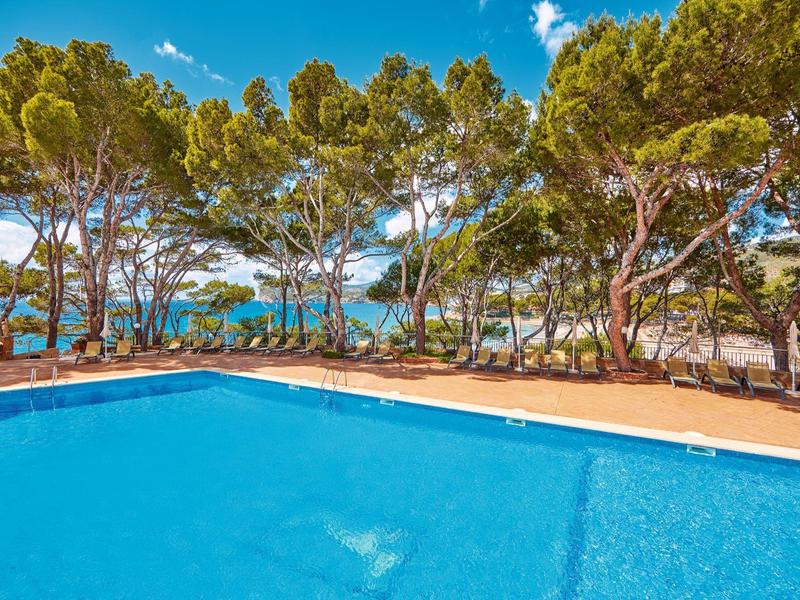 Duży basen z czystą wodą, otoczony drzewami, z widokiem na morze pod błękitnym niebem.