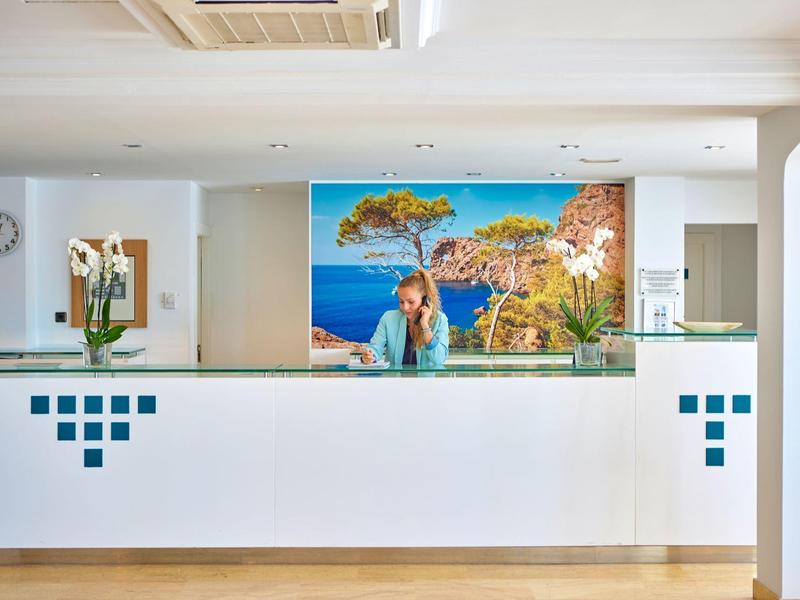 Nowoczesna recepcja hotelowa z białym kontuarem i muralem przedstawiającym letni krajobraz nad morzem.