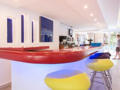 Moderner Barbereich mit roter Theke, weißen Wänden und gelben Barhockern, helle Raumbeleuchtung.