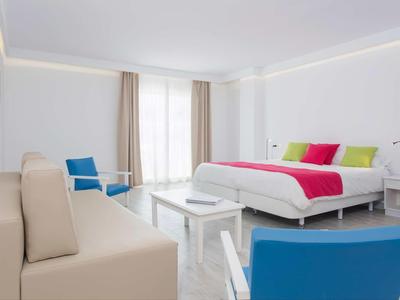 Helles Hotelzimmer mit großem Bett, blauen Sesseln, beige Couch und weißen Möbeln.