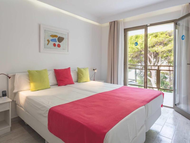 Helles Schlafzimmer mit Doppelbett, bunten Kissen, beigem Bettüberwurf und Balkon mit Naturblick.