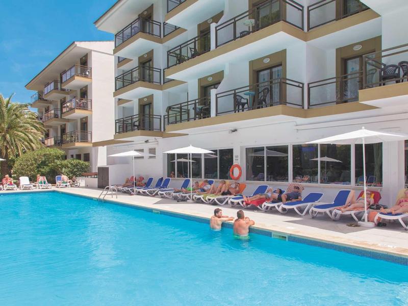 Großer Pool vor mehrstöckigem Hotel mit Liegestühlen und Menschen im Wasser bei blauem Himmel.