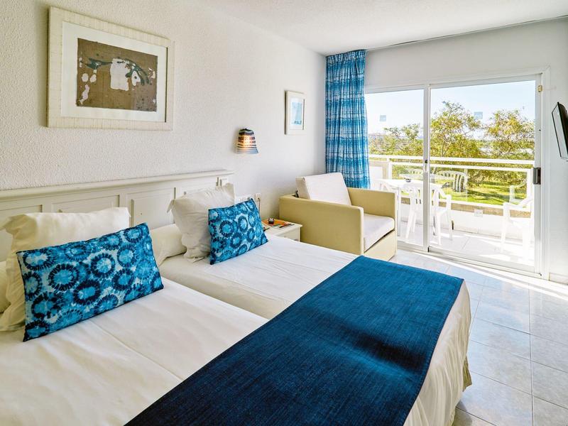 Camera d'hotel luminosa con letto matrimoniale, accenti blu e balcone con vista sul verde.