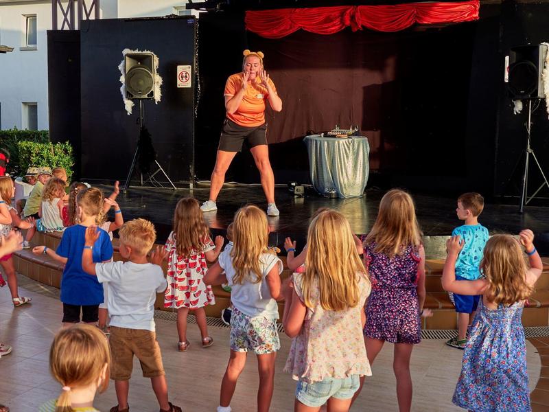 Bambini seguono i movimenti di un'istruttrice di danza sul palco durante un'attività all'aperto.