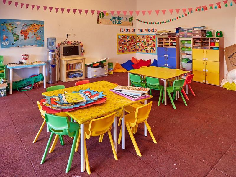 Aula di asilo colorata con piccoli tavoli, sedie e materiali didattici ludici.