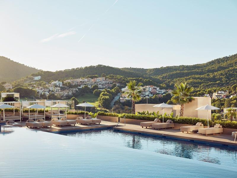 Großer Hotelpool mit Sonnenliegen und Bergen im Hintergrund bei klarem Himmel.