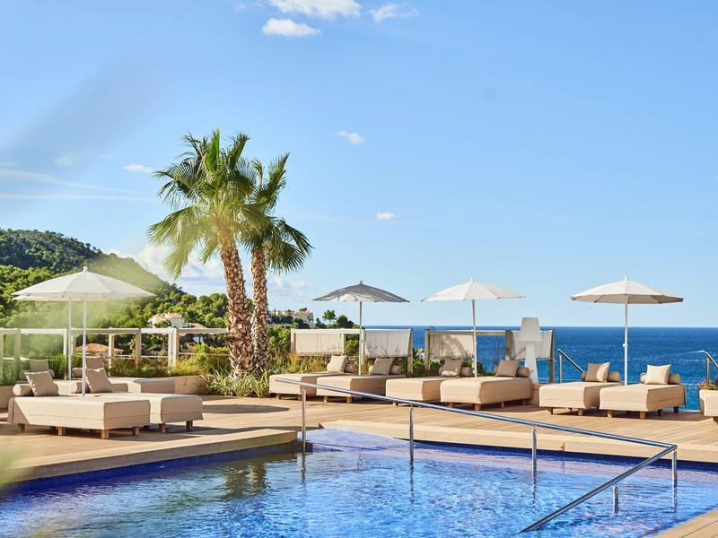 Moderner Hotelpool mit Sonnenliegen, Sonnenschirmen und Blick auf das Meer unter blauem Himmel.