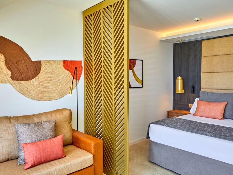 Modernes Hotelzimmer mit Doppelbett, Sofa und dekorativem Raumteiler in warmen Farben.