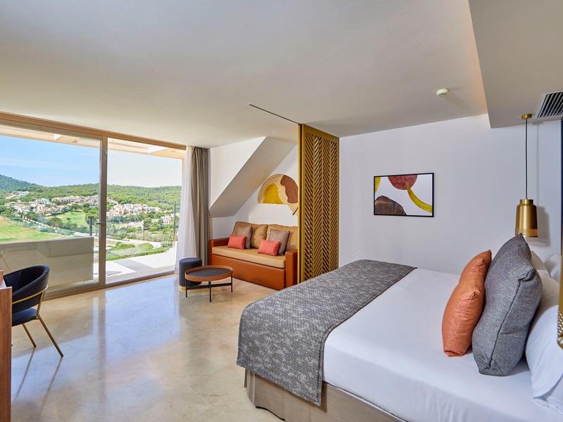 Modernes Hotelzimmer mit Doppelbett, Sofa, großem Fenster und Blick auf grüne Landschaft.