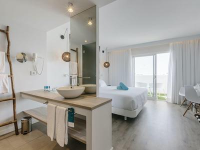 Habitación de hotel moderna con cama doble, paredes claras, gran ventana y baño abierto.