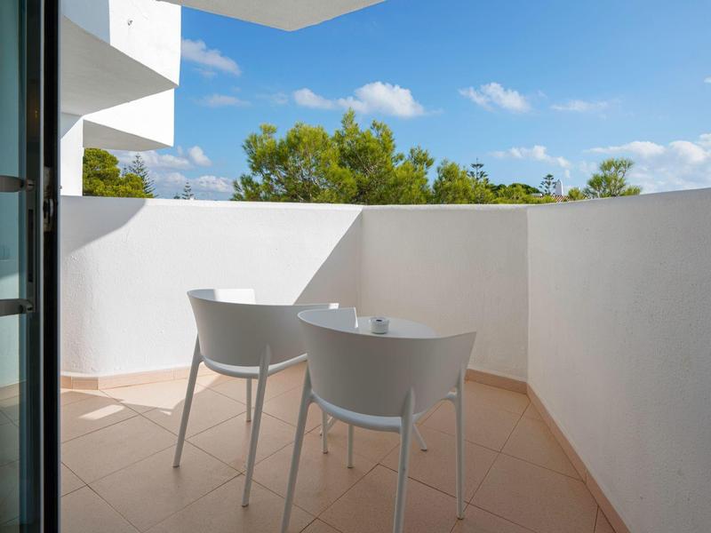 Balcón con dos sillas grises y mesa, vista a árboles verdes y cielo azul.