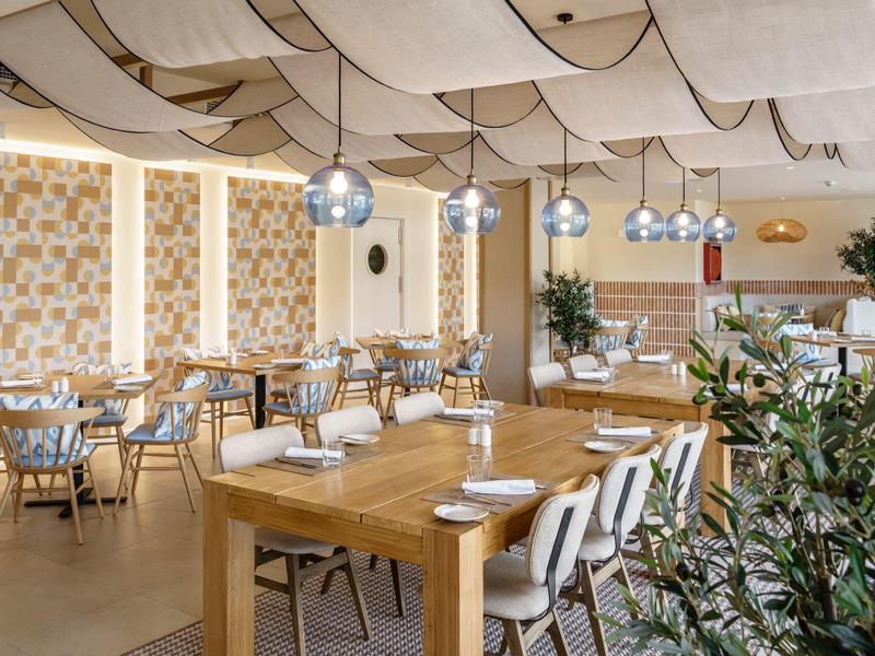 Restaurante de hotel moderno y luminoso con mesas de madera, sillas y plantas decorativas.