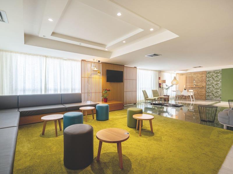 Sala común moderna con alfombra verde, taburetes y grandes ventanas que permiten mucha luz.