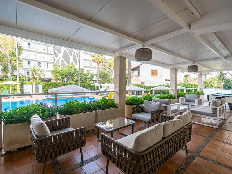 Terraza cubierta con muebles de mimbre y vista a una piscina junto a edificios del hotel.